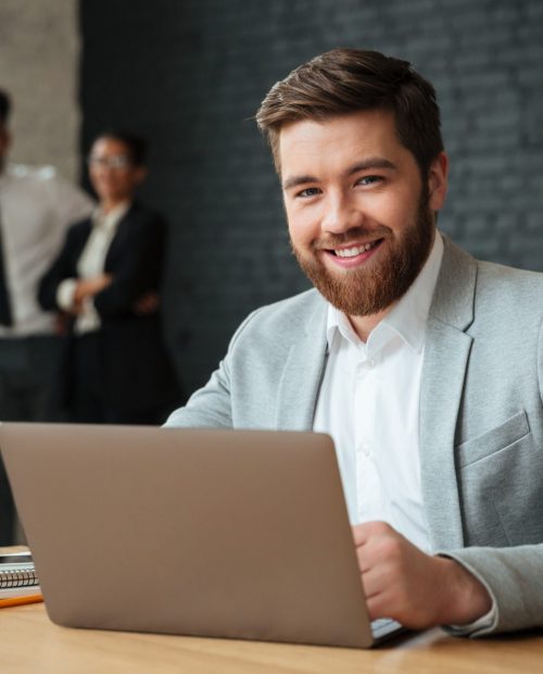 Image of cheerful young caucasian businessman sitting indoors using laptop computer. Looking camera.